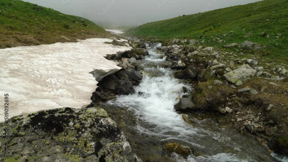 Clean water cascading down the stone bed. Slow motion of a mountain river. Snow lies along the river bed and green grass grows. In the background of the mountain in the fog. Wild nature Clean water cascading down the stone bed. Slow motion of a mountain river. Snow lies along the river bed and green grass grows. In the background of the mountain in the fog. Wild nature