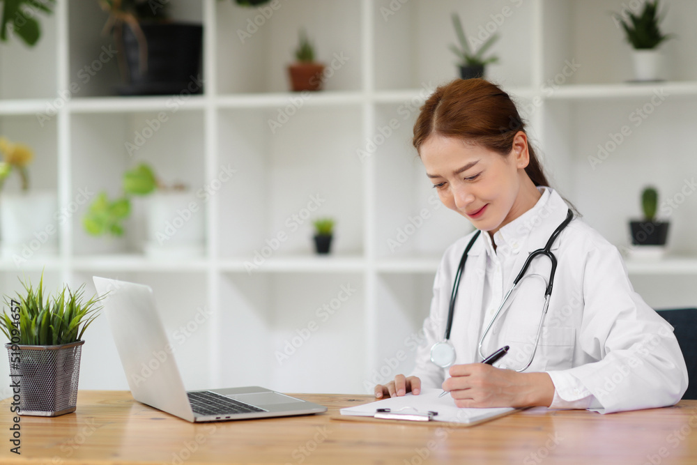 Beautiful Asian woman doctor is taking notes looking at computer screen in her office. Concept of working with information to give the right diagnosis. Beautiful Asian woman doctor is taking notes looking at computer screen in her office. Concept of working with information to give the right diagnosis.