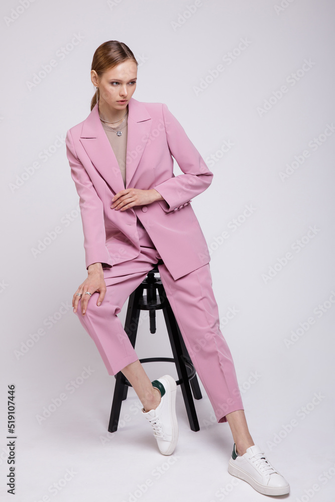 Fashion photo of a beautiful elegant young woman in a pretty soft pink oversized suit, jacket, pants, sneakers posing over white, soft gray background. Studio Shot.