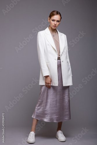 Fashion photo of a beautiful elegant young woman in a pretty white jacket, long metallic silver platinum skirt, sneakers posing gray background. Studio Shot. Portrait