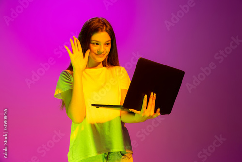 Portrait of young girl, student having online meeting on laptop isolated over pink background in neon light