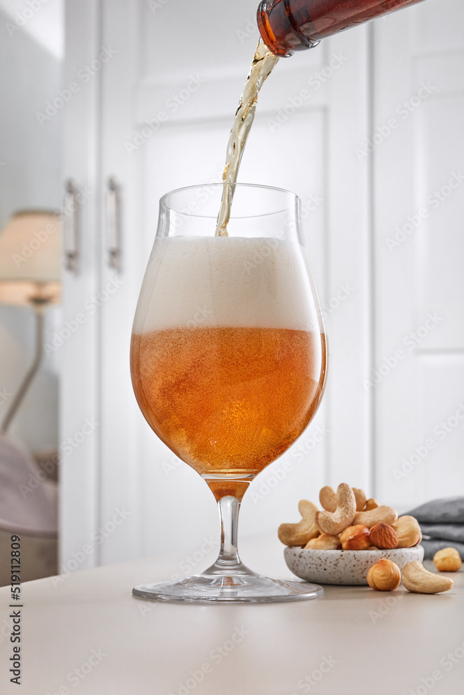 Glass of light beer and snack in a home interior. Light lager beer is pouring into a mug from a bottle on a blurred background of a home interior, a wardrobe and a lamp. Selective focus Glass of light beer and snack in a home interior. Light lager beer is pouring into a mug from a bottle on a blurred background of a home interior, a wardrobe and a lamp. Selective focus
