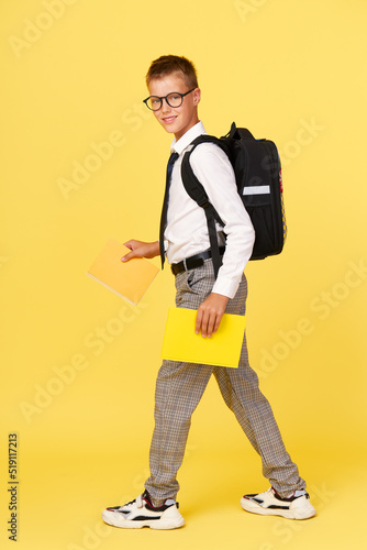 Portrait of a schoolboy in glasses with textbooks and a backpack on a yellow background steps to the left. Back to school