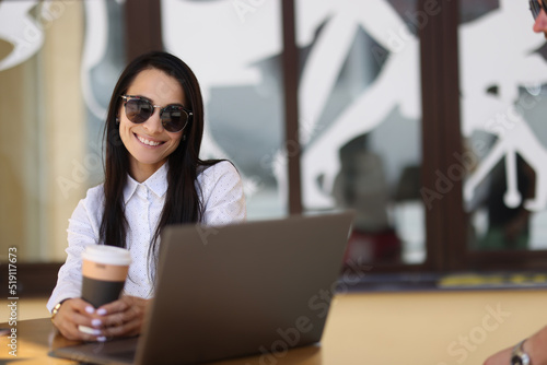 Smiling sun woman holding coffee and working on laptop in cafe