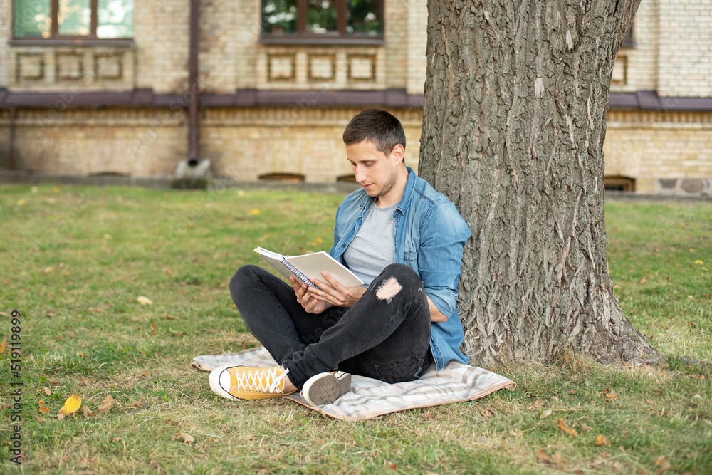 Young man relaxing and reading book while sit on lawn. Happy Man sitting On Green Grass and study remotely. Adult student reading in the park. Enjoying student life. smiling man working on outdoor Young man relaxing and reading book while sit on lawn. Happy Man sitting On Green Grass and study remotely. Adult student reading in the park. Enjoying student life. smiling man working on outdoor