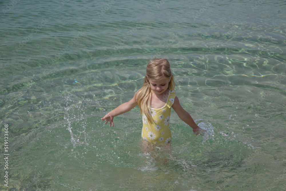 Adorable toddler girl in yellow swimsuit splashing water at the beach. Idyllic summer vacation concept.