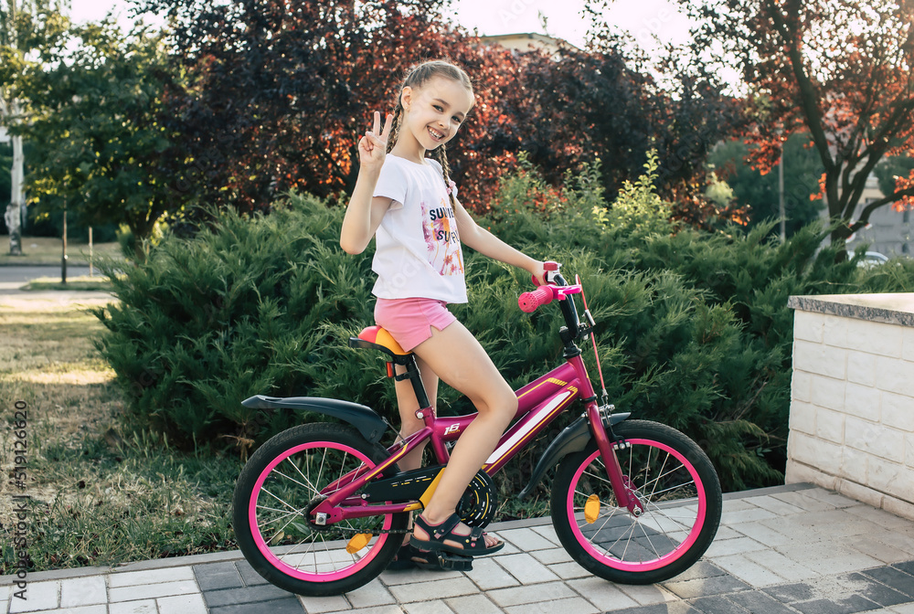 Girl child is dressed in summer clothes and rides a bicycle. Happy smiling child sits on a bike and shows victoria gesture. Girl with pigtails smiling Girl child is dressed in summer clothes and rides a bicycle. Happy smiling child sits on a bike and shows victoria gesture. Girl with pigtails smiling