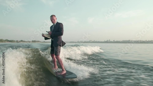 A surfer jumping on a wakeboard in a suit with a book in his hands takes photos of the pages of the book with his phone. An experienced wakeboarder sprays water drops into the camera. photo