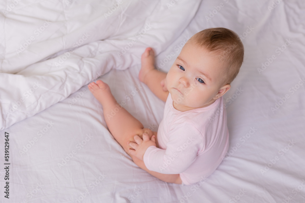 baby in a pink bodysuit is sitting with her back to the camera in a crib on snow-white bed linen