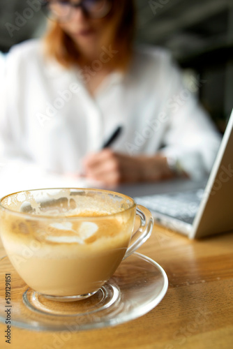 Beautiful business woman making notes on the background and transparent cup of coffee and laptop on the foreground photo