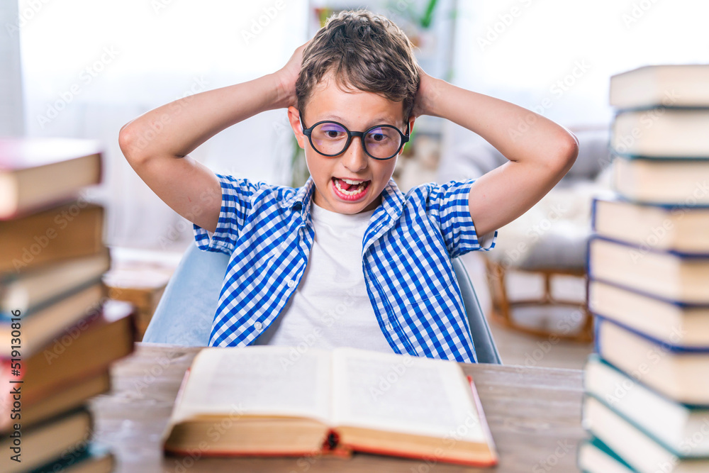 schoolboy tired of learning, wearing glasses, doing homework, sitting indoors between stacks of books. child screams emotionally, indignant because of large amount of homework. Doesn't want to study. schoolboy tired of learning, wearing glasses, doing homework, sitting indoors between stacks of books. child screams emotionally, indignant because of large amount of homework. Doesn't want to study.