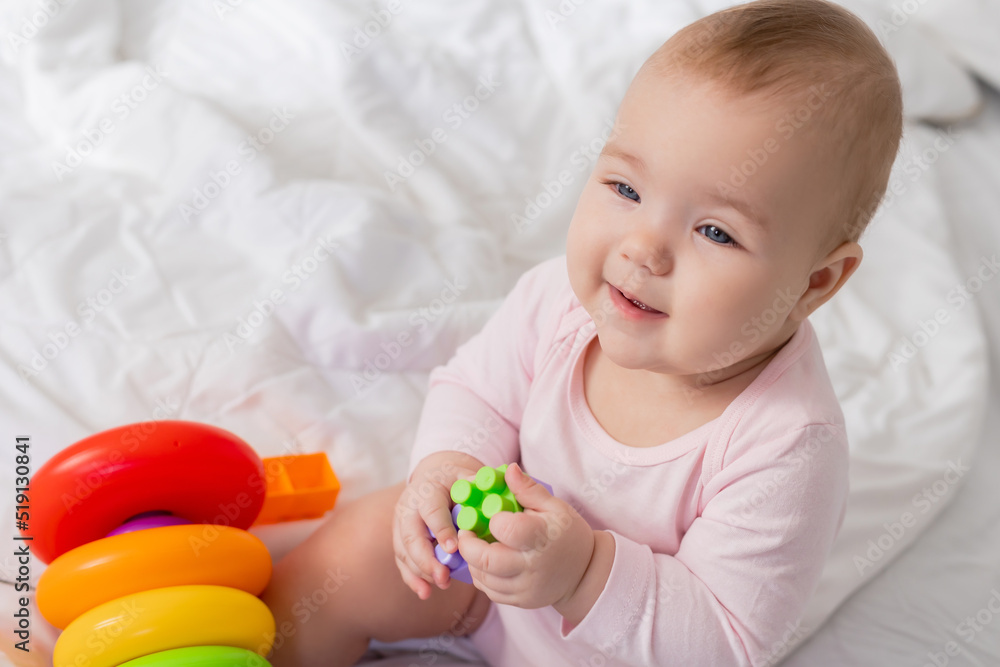 baby in a pink bodysuit plays with an educational multicolored pyramid while sitting in bed baby in a pink bodysuit plays with an educational multicolored pyramid while sitting in bed