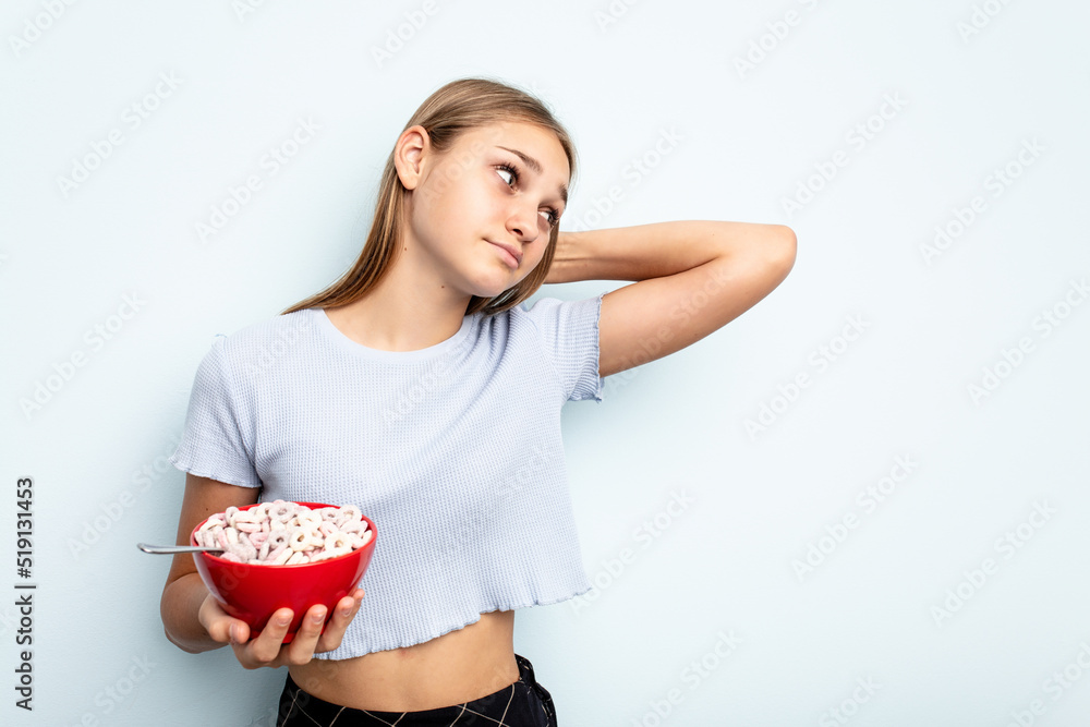Young caucasian girl eating cereals isolated on blue background touching back of head, thinking and making a choice. Young caucasian girl eating cereals isolated on blue background touching back of head, thinking and making a choice.