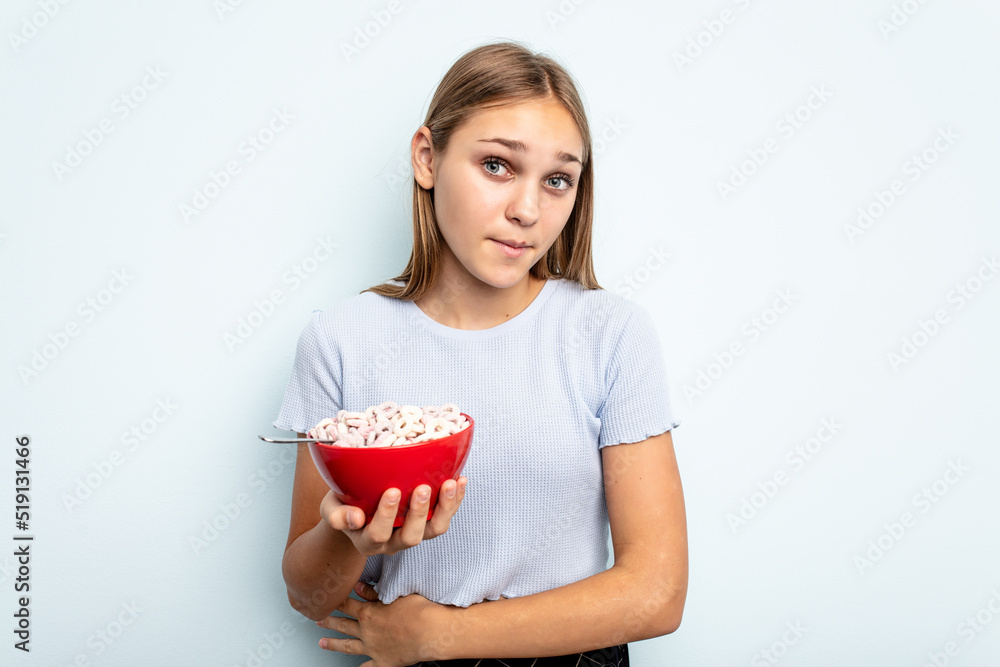 Young caucasian girl eating cereals isolated on blue background shrugs shoulders and open eyes confused. Young caucasian girl eating cereals isolated on blue background shrugs shoulders and open eyes confused.