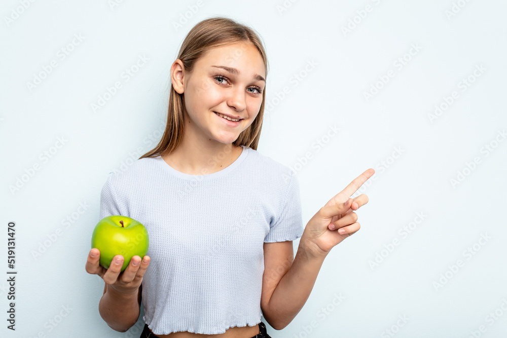 Young caucasian girl holding an apple isolated on blue background smiling and pointing aside, showing something at blank space. Young caucasian girl holding an apple isolated on blue background smiling and pointing aside, showing something at blank space.