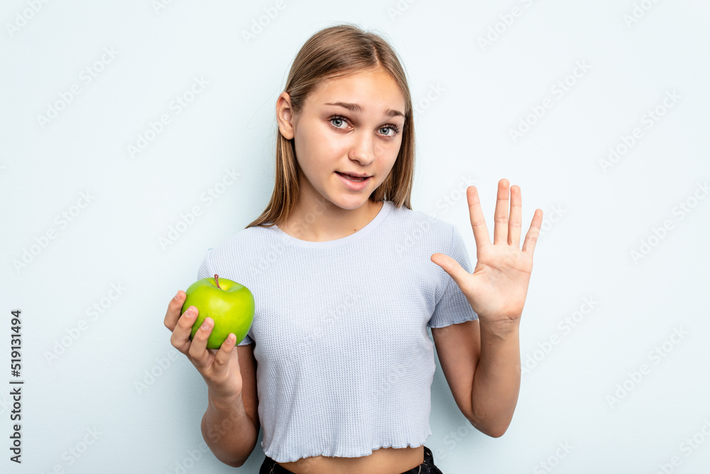 Young caucasian girl holding an apple isolated on blue background smiling cheerful showing number five with fingers. Young caucasian girl holding an apple isolated on blue background smiling cheerful showing number five with fingers.