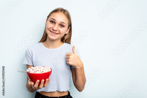Young caucasian girl eating cereals isolated on blue background smiling and raising thumb up