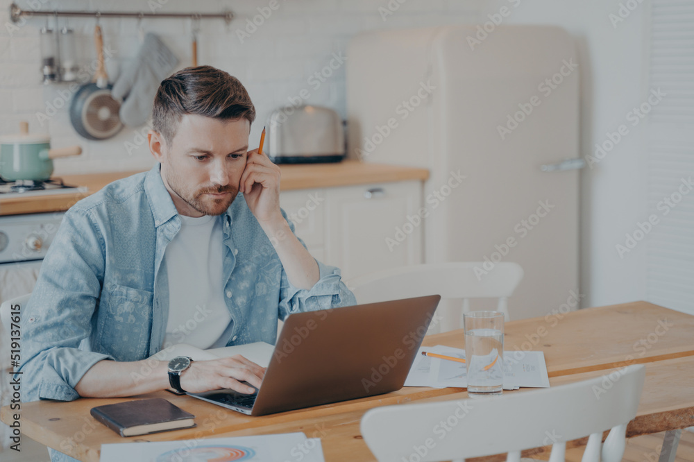 Serious focused bearded businessman in casual clothes working on laptop while sitting at kitchen table