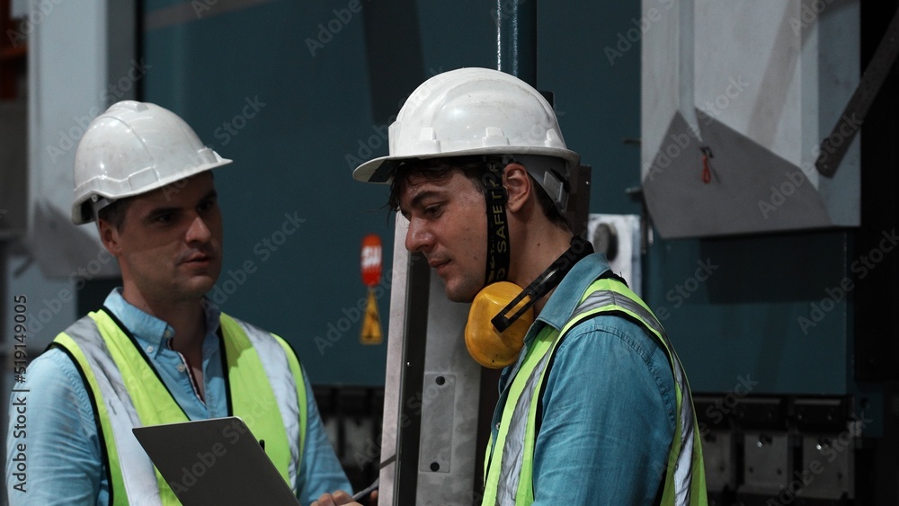 Close up two specialists inspect commercial, Wear safety uniform and White helmet work on laptop computer and clipboard. Two American people technician and Worker talk on a radio meet in a factory. Close up two specialists inspect commercial, Wear safety uniform and White helmet work on laptop computer and clipboard. Two American people technician and Worker talk on a radio meet in a factory.