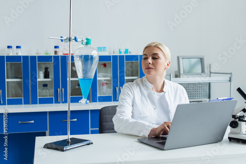 Scientist using laptop and looking at flask with liquid in laboratory.