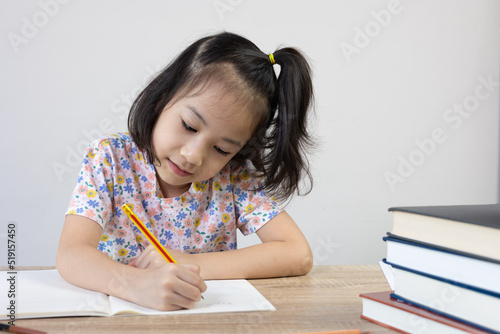 Adorable asian girl is studying her lesson on the table with a lot of books and pencil. It shows concept of smart and clever child learning by herself to gain knowledge which is basic education.