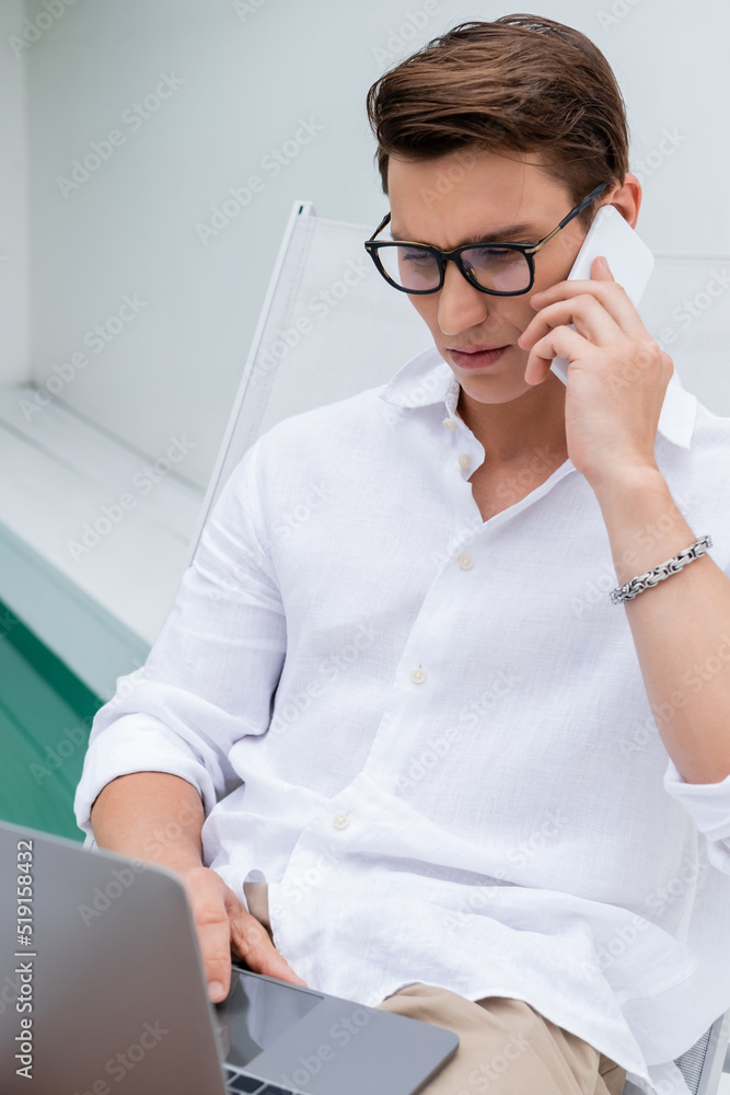 focused freelancer in white shirt and eyeglasses using laptop and talking on cellphone outdoors.
