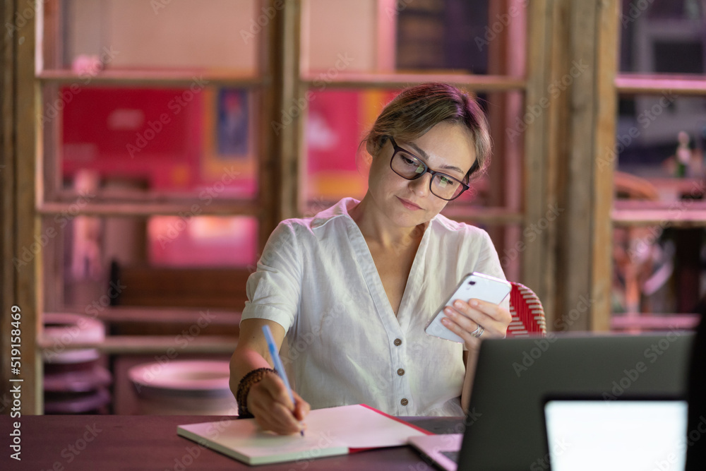 Focused woman in eyeglasses looking at smartphone writing down notes, studying online in cafe, using laptop. Concentrated female language tutor preparing for virtual lesson. Distance education Focused woman in eyeglasses looking at smartphone writing down notes, studying online in cafe, using laptop. Concentrated female language tutor preparing for virtual lesson. Distance education