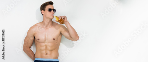 man with muscular torso standing near white wall in sunglasses and drinking beer, banner.