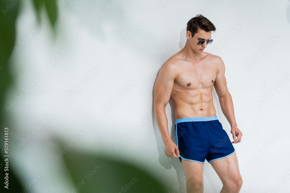 Obraz premium shirtless man in sunglasses and swimming trunks near white wall on blurred foreground.