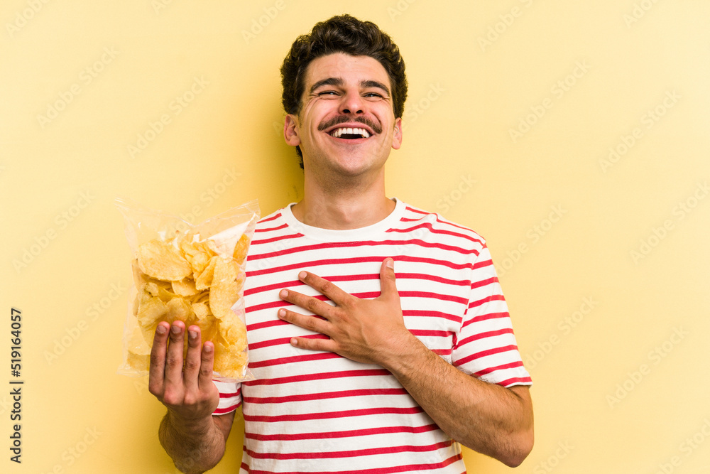 Young caucasian man holding a bag of chips isolated on yellow background laughs out loudly keeping hand on chest. Young caucasian man holding a bag of chips isolated on yellow background laughs out loudly keeping hand on chest.