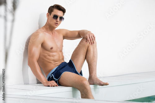 man with muscular torso sitting near pool in sunglasses and swimming trunks. photo