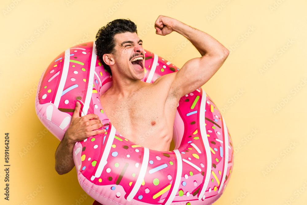 Young caucasian man holding an inflatable donut isolated on yellow background raising fist after a victory, winner concept.