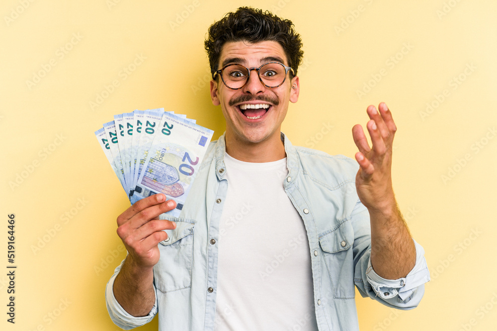 Young caucasian man holding banknotes isolated on yellow background receiving a pleasant surprise, excited and raising hands. Young caucasian man holding banknotes isolated on yellow background receiving a pleasant surprise, excited and raising hands.