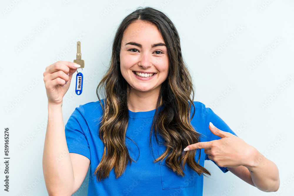 Young caucasian woman holding home keys isolated on blue background person pointing by hand to a shirt copy space, proud and confident Young caucasian woman holding home keys isolated on blue background person pointing by hand to a shirt copy space, proud and confident