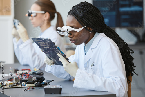 Portrait of young black woman wearing magnifying glasses and inspecting electronic parts in quality control lab photo