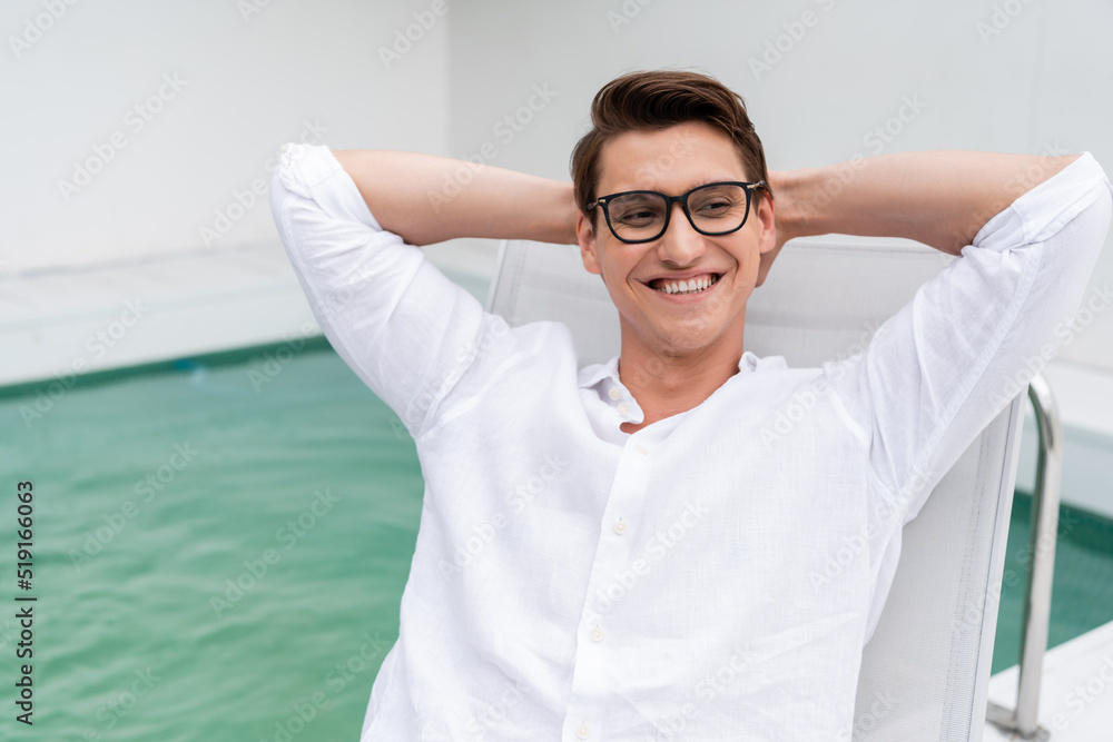 joyful man in eyeglasses looking away while resting with hands behind head near pool. joyful man in eyeglasses looking away while resting with hands behind head near pool.