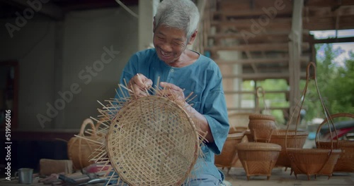 Slow motion scene of a happy smiling Asian male farmer who is an elderly native basketry artisan weave basket from bamboo that are cut into strips for weaving folk crafts in rural areas of Thailand.