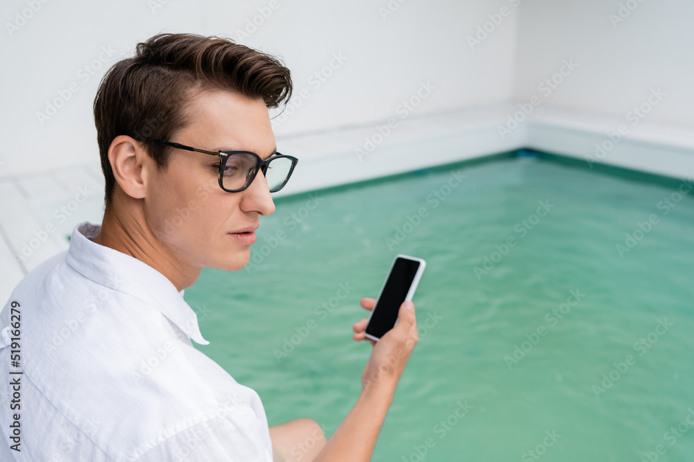 man in eyeglasses holding smartphone with blank screen near blurred pool.