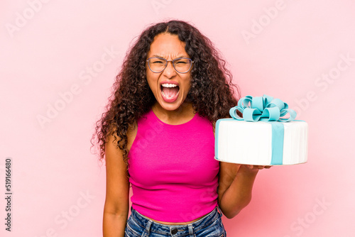 Young caucasian woman holding a cake isolated on pink background screaming very angry and aggressive.