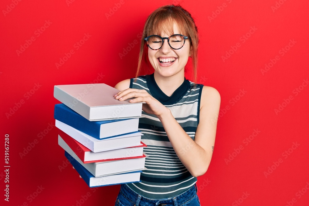 Redhead young woman holding a pile of books celebrating crazy and amazed for success with open eyes screaming excited. Redhead young woman holding a pile of books celebrating crazy and amazed for success with open eyes screaming excited.
