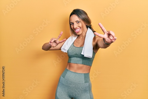 Beautiful hispanic woman wearing sportswear and towel smiling with tongue out showing fingers of both hands doing victory sign. number two.