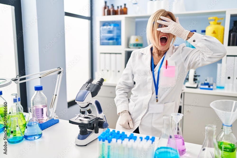 Middle age blonde woman working at scientist laboratory peeking in shock covering face and eyes with hand, looking through fingers with embarrassed expression. Middle age blonde woman working at scientist laboratory peeking in shock covering face and eyes with hand, looking through fingers with embarrassed expression.