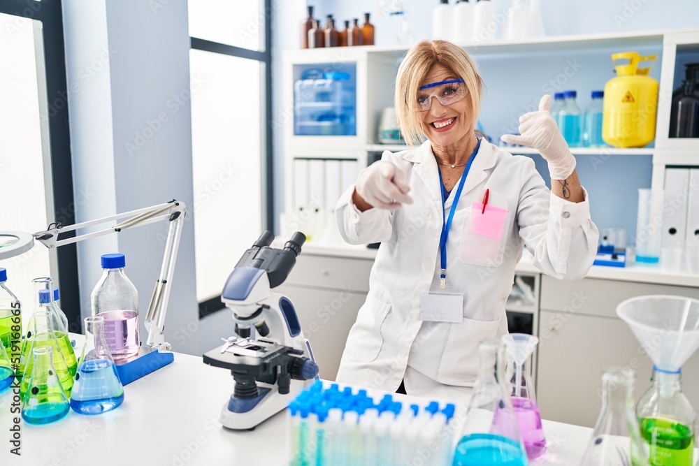Middle age blonde woman working at scientist laboratory smiling doing talking on the telephone gesture and pointing to you. call me. Middle age blonde woman working at scientist laboratory smiling doing talking on the telephone gesture and pointing to you. call me.