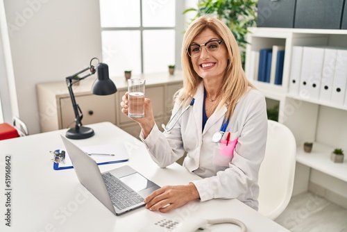 Middle age blonde doctor woman working using computer laptop drinking water looking positive and happy standing and smiling with a confident smile showing teeth