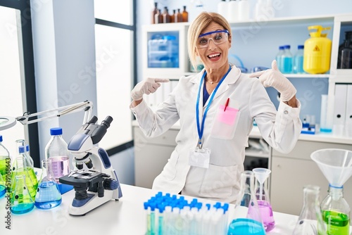 Middle age blonde woman working at scientist laboratory looking confident with smile on face  pointing oneself with fingers proud and happy.