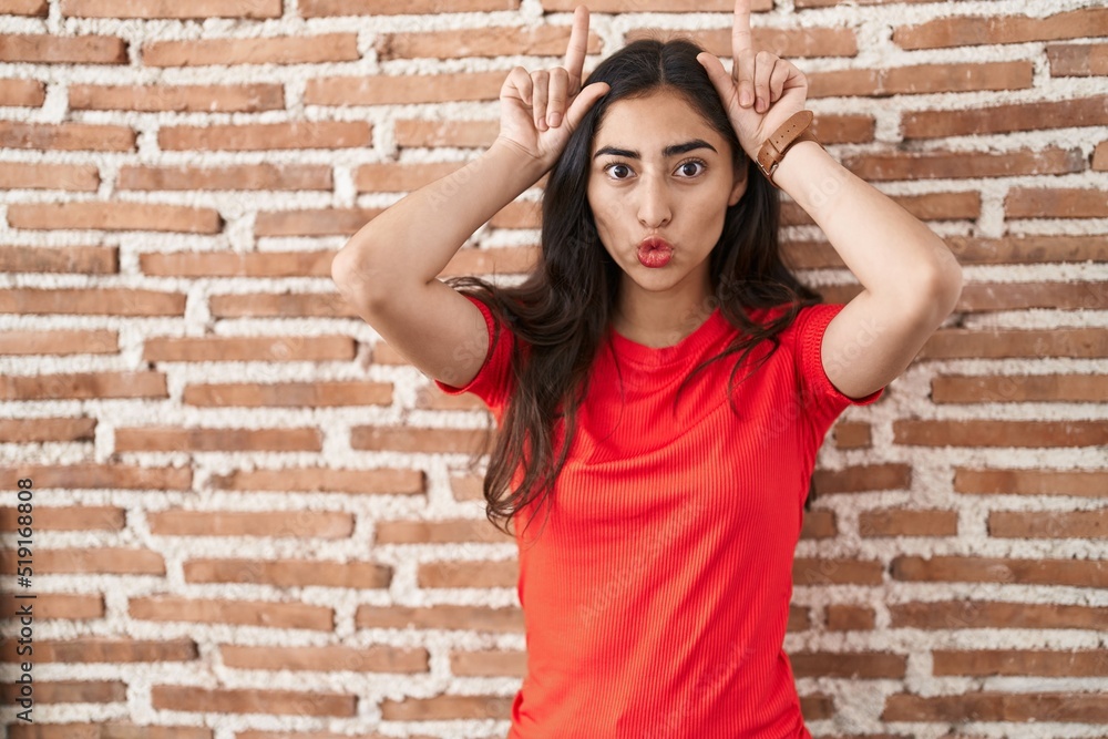 Young teenager girl standing over bricks wall doing funny gesture with finger over head as bull horns Young teenager girl standing over bricks wall doing funny gesture with finger over head as bull horns