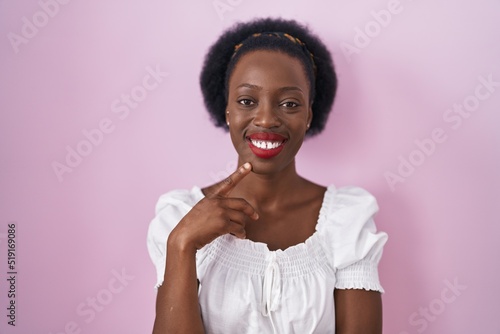African woman with curly hair standing over pink background with hand on chin thinking about question, pensive expression. smiling and thoughtful face. doubt concept.