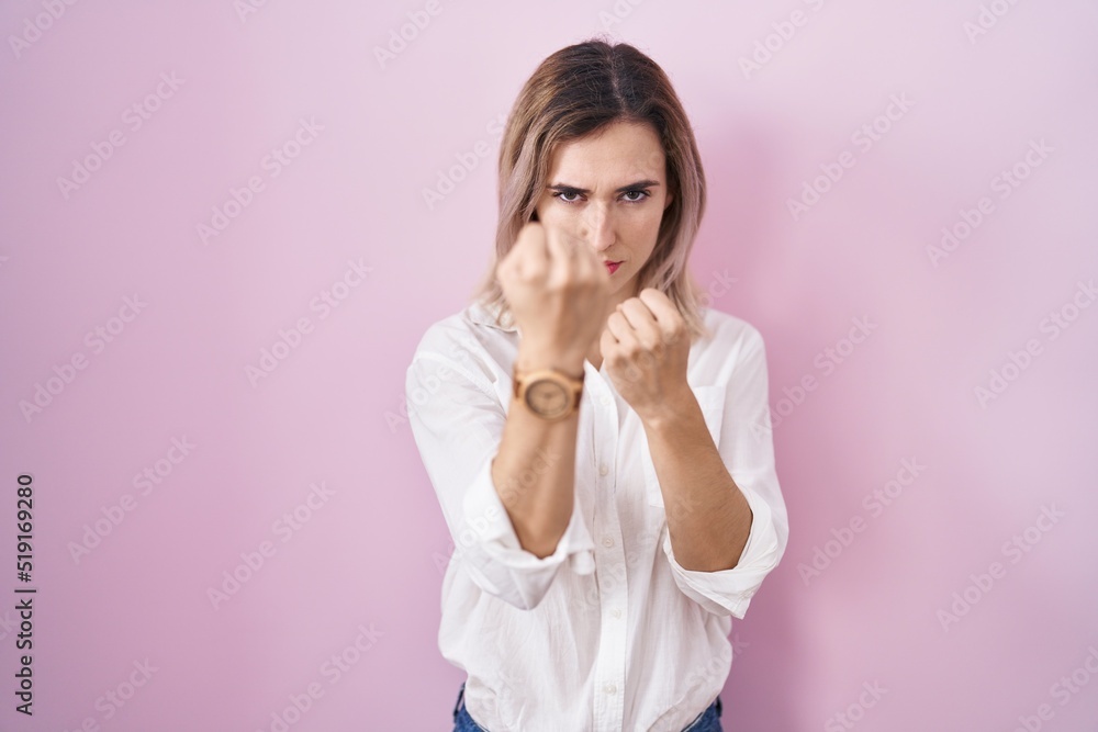 Young beautiful woman standing over pink background ready to fight with fist defense gesture, angry and upset face, afraid of problem Young beautiful woman standing over pink background ready to fight with fist defense gesture, angry and upset face, afraid of problem