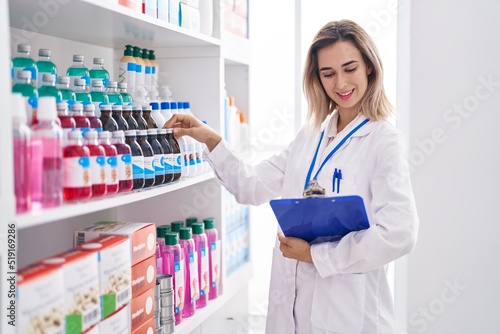 Young woman pharmacist smiling confident holding checklist and medicine bottle at pharmacy