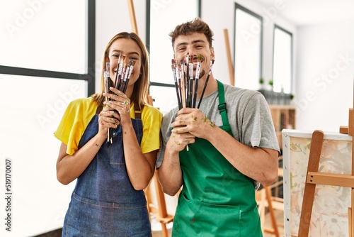 Young hispanic artist couple smiling happy covering mouth with paintbrushes at art studio.